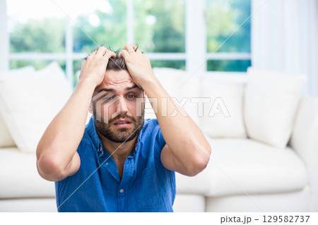 Man gripping hair, sitting on floor in front of white sofa cushions under large multi-pane window 129582737