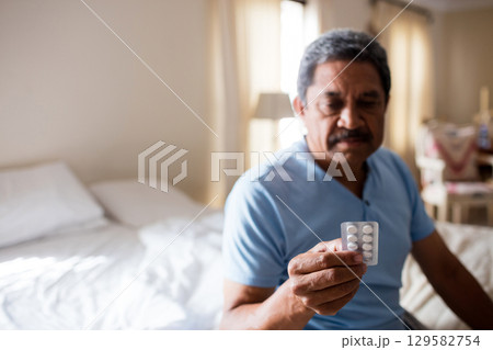 Senior man sitting on bed edge in bedroom examining blister pack of white pills, copy space Senior man sitting on bed edge in bedroom examining blister pack of white pills, copy space 129582754