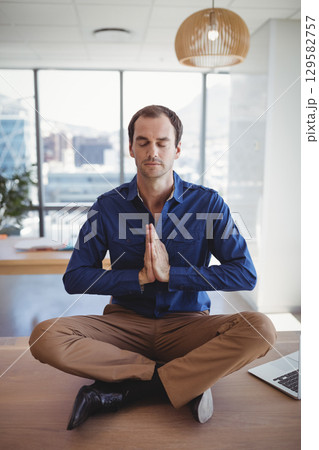 Man meditating at office on wooden desk under pendant lamp near laptop papers and plant Man meditating at office on wooden desk under pendant lamp near laptop papers and plant 129582757