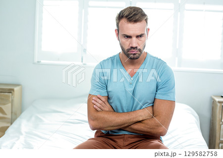 Mid-adult man sitting on bed edge in bright bedroom crossing arms near nightstand, box and blinds Mid-adult man sitting on bed edge in bright bedroom crossing arms near nightstand, box and blinds 129582758