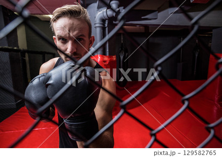 Male athlete holding boxing gloves up inside chain-link fight cage at gym with red floor 129582765