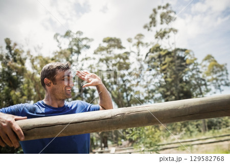 Middle-aged man leaning on wooden beam at obstacle course, shielding eyes in blue shirt, copy space 129582768