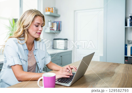 Woman in her twenties typing on laptop at table in kitchen with pink mug, copy space Woman in her twenties typing on laptop at table in kitchen with pink mug, copy space 129582793
