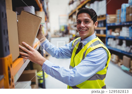 Man standing in warehouse aisle wearing hi-vis safety vest holding cardboard box, copy space 129582813