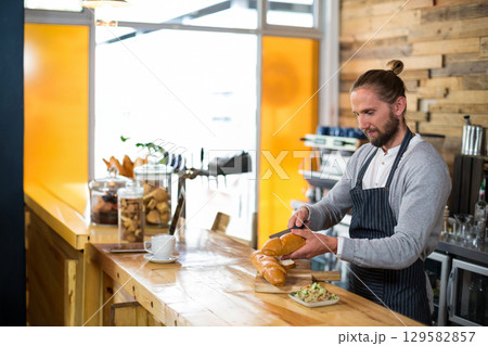 Male barista slicing braided loaf on cutting board at cafe counter with glass jars, copy space Male barista slicing braided loaf on cutting board at cafe counter with glass jars, copy space 129582857