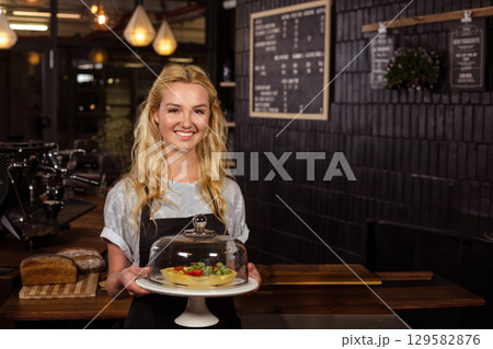 Female barista wearing apron standing behind espresso machine at cafe holding tart under glass dome 129582876