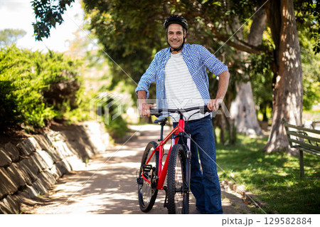 Man wearing helmet pushing red bicycle along sunlit park path with stone wall and park bench Man wearing helmet pushing red bicycle along sunlit park path with stone wall and park bench 129582884
