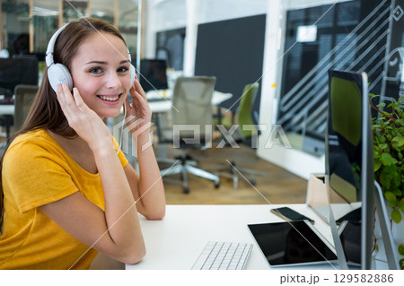 Adjusting over-ear headphones while sitting at desk in workspace with computer and smartphone 129582886