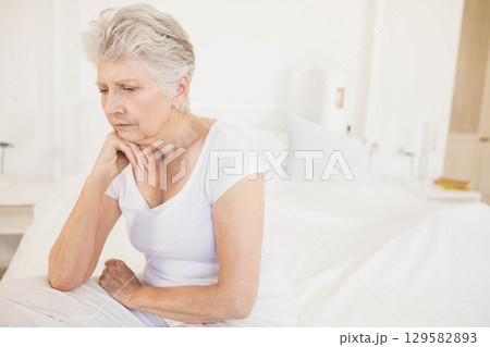 Senior woman sitting on bed edge in bedroom leaning on hand gazing at book, copy space Senior woman sitting on bed edge in bedroom leaning on hand gazing at book, copy space 129582893