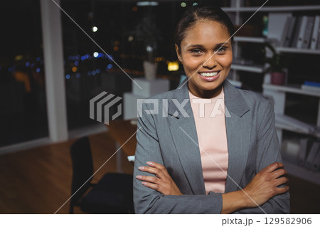 Businesswoman standing in modern office at night with desk, shelves with plant and city skyline Businesswoman standing in modern office at night with desk, shelves with plant and city skyline 129582906