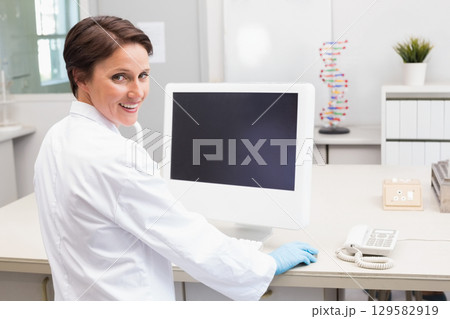 Female researcher wearing lab coat and gloves sitting at lab bench smiling by computer DNA model Female researcher wearing lab coat and gloves sitting at lab bench smiling by computer DNA model 129582919