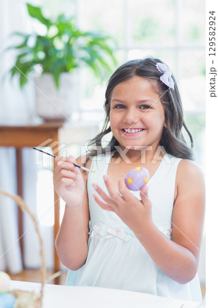 School-age girl painting egg at table by bright window at home with wicker basket and paintbrush 129582924