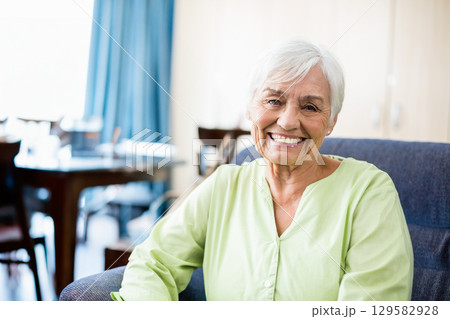 Senior woman sitting in armchair at home by dining table set, smiling at camera, copy space 129582928