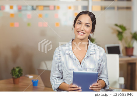 Female professional wearing blouse smiling while holding tablet at desk in office with sticky notes Female professional wearing blouse smiling while holding tablet at desk in office with sticky notes 129582940