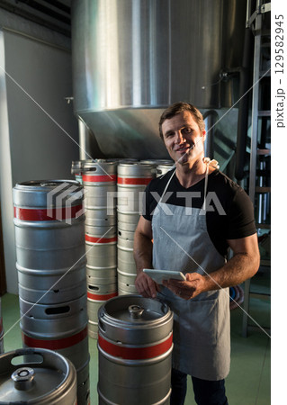 Senior man wearing apron and holding tablet by stainless fermenter tank among kegs in brewery 129582945