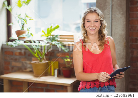 Teenage girl standing in studio holding tablet by large window over bench with plants, copy space 129582961