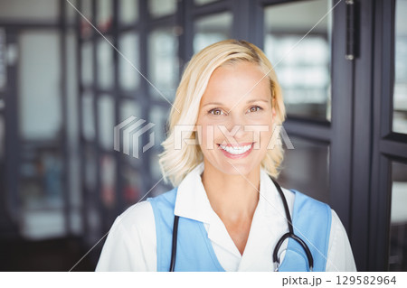 Female nurse in thirties standing in medical corridor wearing light blue uniform and stethoscope 129582964