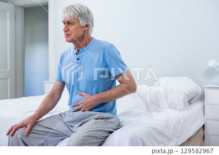 Senior man clutching abdomen while sitting on bed edge in tidy bedroom with nightstand lamp Senior man clutching abdomen while sitting on bed edge in tidy bedroom with nightstand lamp 129582967