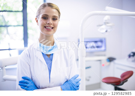 Female dentist standing with arms crossed in dental clinic showing dental chair and instrument tray 129582985