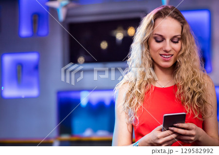 Woman in red sleeveless top holding smartphone standing by blue LED shelves in lounge, copy space 129582992