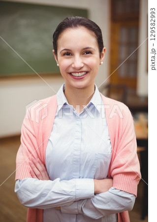 Smiling female teacher wearing blue shirt pink cardigan classroom chalkboard, desks, chairs, door 129582993