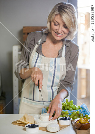 Mature woman slicing soft cheese with cheese knife on cutting board at kitchen counter with grapes Mature woman slicing soft cheese with cheese knife on cutting board at kitchen counter with grapes 129582997