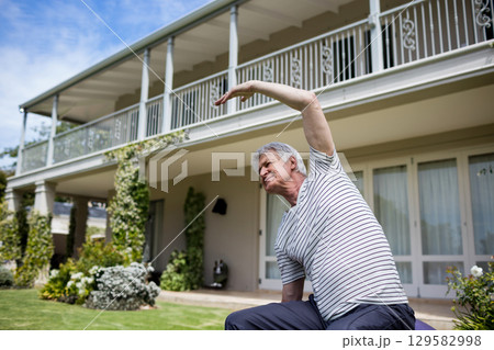 Senior man balancing on exercise ball stretching sideways on lawn by porch pillars, copy space 129582998