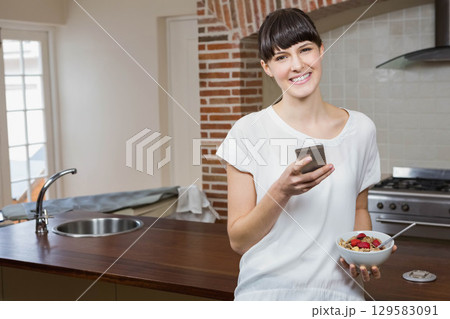 Woman in her twenties leaning on countertop holding smartphone and cereal in kitchen, copy space Woman in her twenties leaning on countertop holding smartphone and cereal in kitchen, copy space 129583091