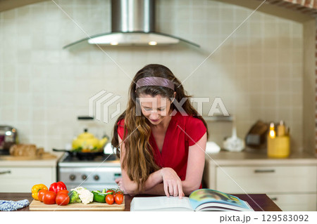 Woman in twenties standing at kitchen island chopping vegetables on cutting board reading cookbook 129583092