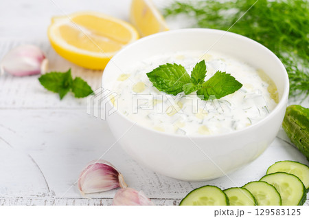 Creamy Greek dip sauce tzatziki in a white bowl garnished with fresh mint, surrounded by cucumber slices and garlic on a rustic wooden table Creamy Greek dip sauce tzatziki in a white bowl garnished with fresh mint, surrounded by cucumber slices and garlic on a rustic wooden table 129583125