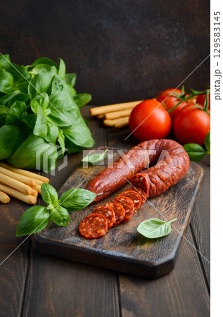 Sliced spicy Chorizo sausage arranged on a wooden board with fresh basil, tomatoes, and breadsticks, creating a vibrant culinary scene Sliced spicy Chorizo sausage arranged on a wooden board with fresh basil, tomatoes, and breadsticks, creating a vibrant culinary scene 129583145