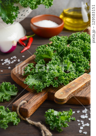 Fresh green kale leaves arranged on wooden cutting board with olive oil, salt, and red chili peppers in a rustic kitchen setting Fresh green kale leaves arranged on wooden cutting board with olive oil, salt, and red chili peppers in a rustic kitchen setting 129583151