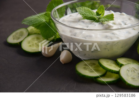 Creamy Greek dip sauce tzatziki in a glass bowl garnished with fresh mint leaves, surrounded by sliced cucumbers and garlic cloves, perfect for appetizers 129583186