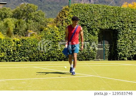 African American man strolling across tennis court carrying racket, sports bag and tennis balls 129583746