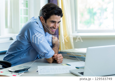 Mid adult man smiling at silver laptop while tapping smartphone then sorting file folders at office 129583749