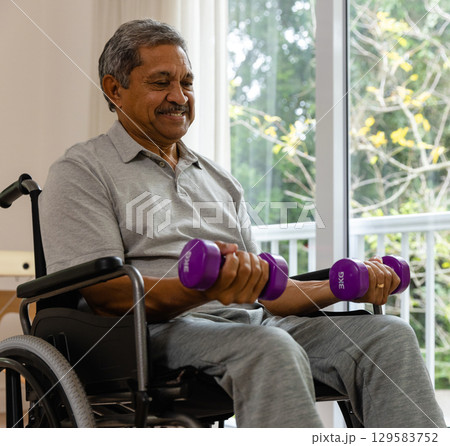 Senior Asian man in wheelchair by glass doors performing curls with purple dumbbells, copy space 129583752