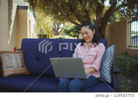 Mature adult Korean woman sitting on blue sofa using silver-gray laptop with pillows, copy space 129583755