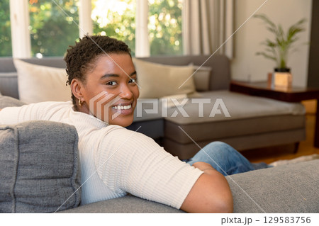 African American adult woman reclining on grey sofa by tall windows at home with folded book 129583756