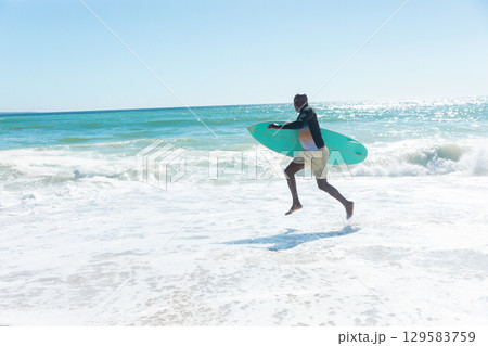 African American man carrying turquoise surfboard while walking on sunlit beach, copy space 129583759