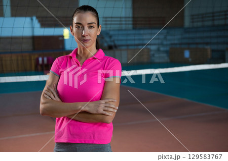 Female volleyball player standing inside gym wearing pink polo and gray leggings by net and benches 129583767