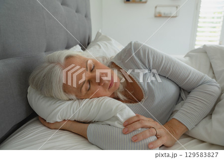 Mature woman lying on side in bedroom showing white pillow comforter grey headboard blinds Mature woman lying on side in bedroom showing white pillow comforter grey headboard blinds 129583827