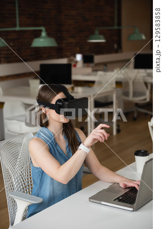 Female employee is wearing VR headset and sitting at white desk, with laptop in open-plan office 129583858