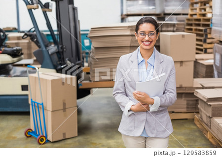 African American woman in blazer smiling and holding folders in warehouse near forklift and dolly 129583859