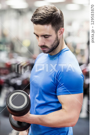 Male weightlifter performing bicep curl with dumbbell at gym wearing blue and black workout attire 129583870