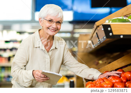 Senior woman inspecting ripe tomatoes on wooden produce bin holding spiral notebook in supermarket 129583871