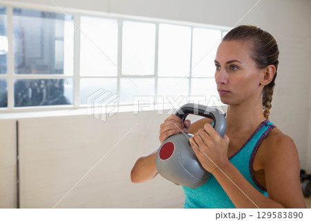 Female athlete holding gray kettlebell with red accent at chest in studio with window, copy space 129583890