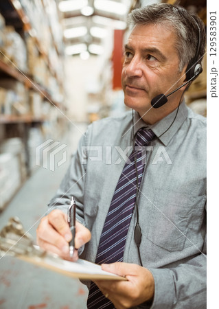 Man wearing tie inspecting boxes in warehouse aisle holding clipboard and headset, copy space Man wearing tie inspecting boxes in warehouse aisle holding clipboard and headset, copy space 129583901
