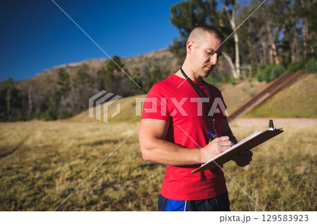 Man standing in grassy field by wooden stairway writing on clipboard with whistle and pen Man standing in grassy field by wooden stairway writing on clipboard with whistle and pen 129583923