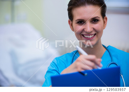 Female nurse smiling filling out chart by bed in ward with clipboard, stethoscope, copy space 129583937