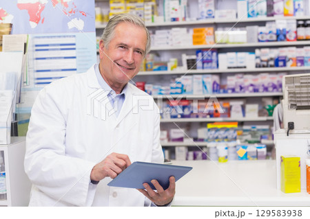 Senior male pharmacist using tablet behind pharmacy counter with brochure stand and medications Senior male pharmacist using tablet behind pharmacy counter with brochure stand and medications 129583938
